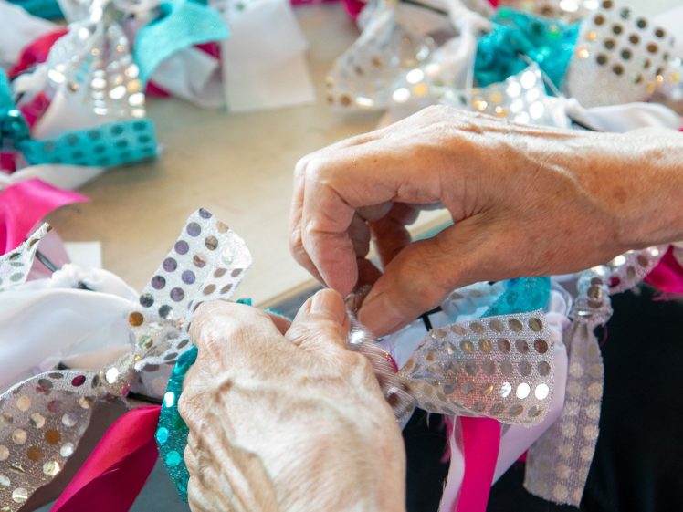 Image of Close-up of hands tying colourful ribbons, including silver with polka dots, pink, and teal, onto a circular base on a table, creating a festive or decorative craft project.