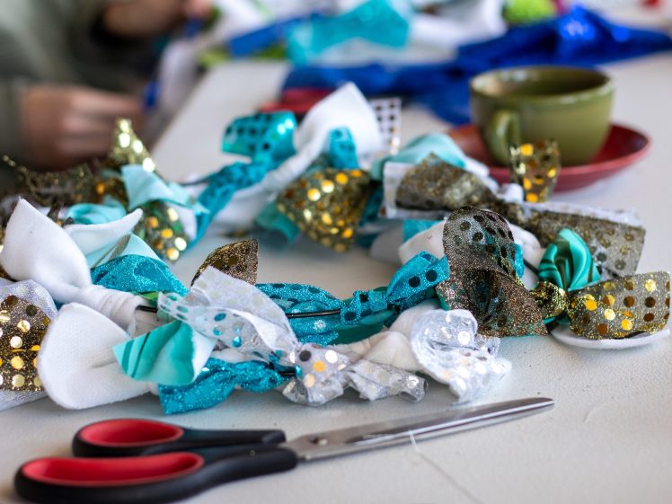 Image of A close-up of a colourful wreath made from blue, white, and gold fabric bows on a table, with scissors, a green cup, and blurred hands working in the background.