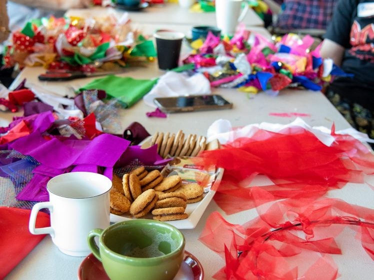 Image of A group of people work on colourful crafts at a table covered with fabric scraps, paper, biscuits, mugs, cups, and saucers. The lively scene suggests a creative, social gathering.