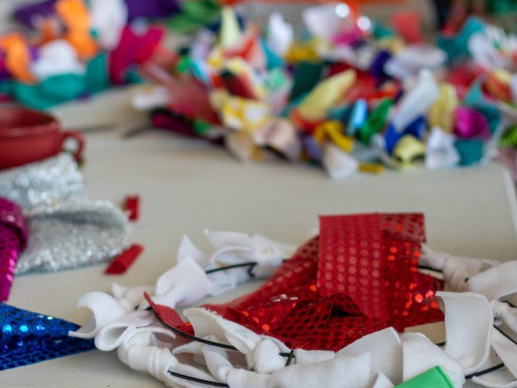 Image of A close-up of a table covered with colourful fabric scraps, including shiny yellow, red, and green pieces, with blurred hands working in the background. The scene suggests a creative crafting or sewing activity.