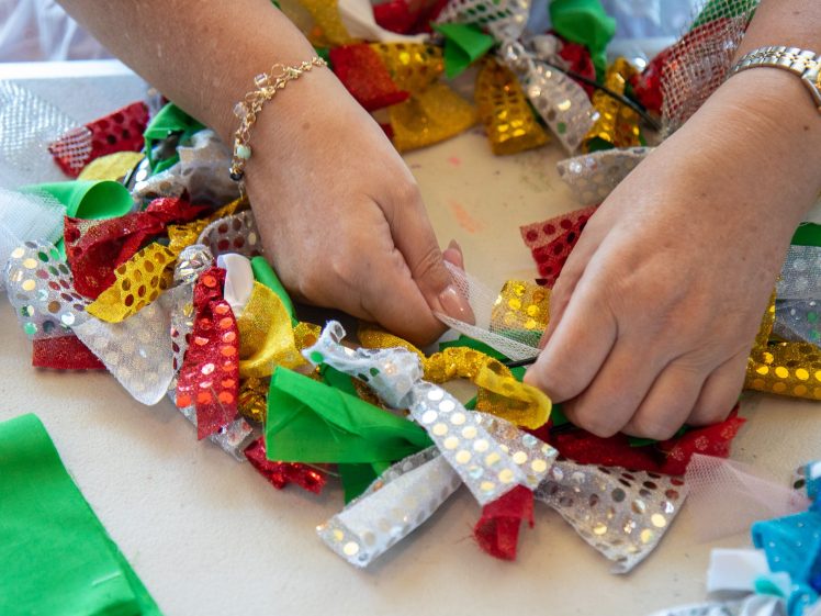 Image of Two hands tie colourful, shiny fabric strips—red, gold, green, and silver—onto a white mesh base, creating a festive wreath. The person wears a gold bracelet and watch.