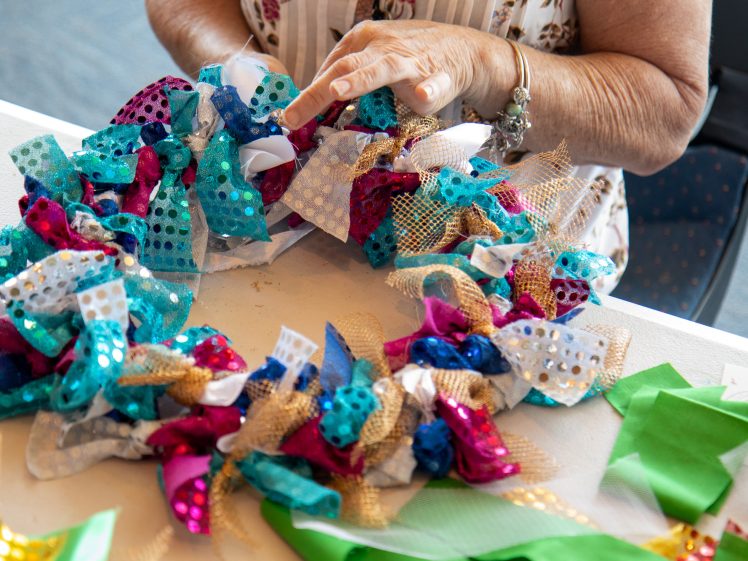 Image of A person’s hands are making a colourful wreath using pieces of blue, pink, gold, and white fabric with sparkly patterns at a table decorated with green ribbon pieces.