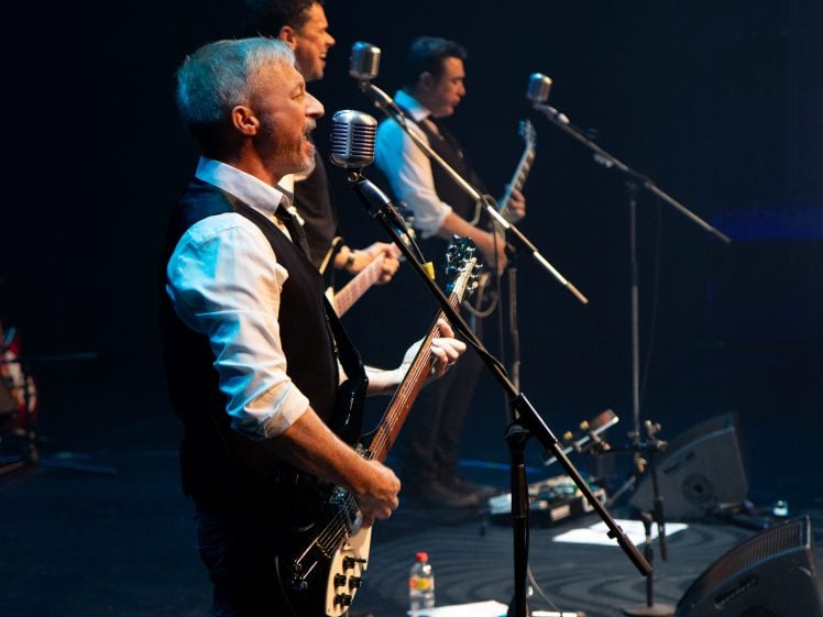 Image of Three male musicians in black waistcoats and white shirts perform on stage with electric guitars and vintage microphones under stage lighting, with musical equipment and cables on the floor.