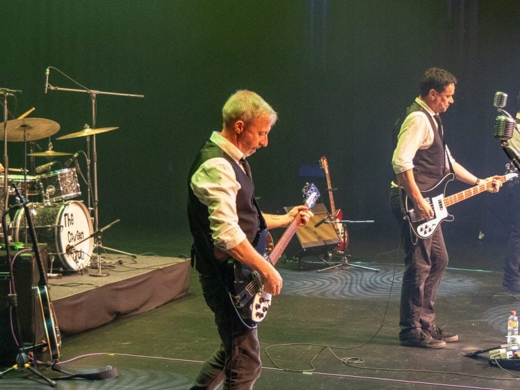 Image of Four male musicians in white shirts and black waistcoats perform on a dimly lit stage with drums, guitars, and vintage microphones. The drummer is at the back, while three others play guitar and sing at the front.