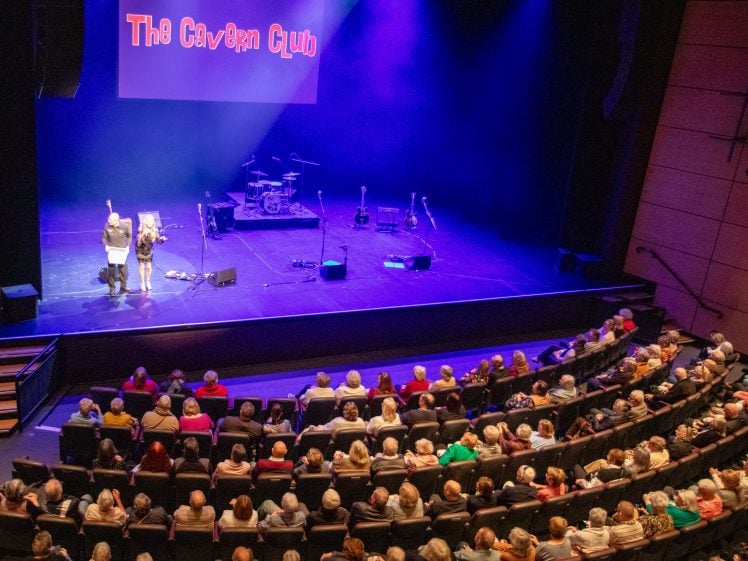 Image of A large audience watches two people speaking on stage under purple lighting, with musical instruments set up and a screen above reading The Cavern Club.