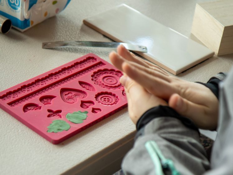 Image of A person shapes dough or clay with their hands next to a pink silicone mould featuring decorative shapes on a table with crafting tools and materials.