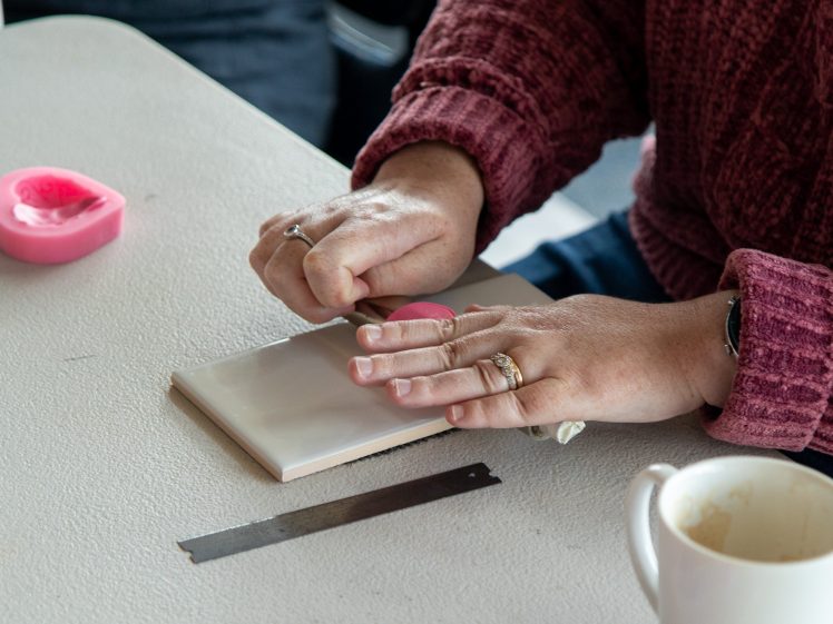 Image of A person in a maroon jumper presses a pink heart-shaped stamp onto a white tile, with a metal blade and a white mug nearby on the table.