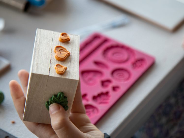 Image of A person holds a wooden block decorated with small orange bird shapes and a green leaf, whilst a pink silicone mould with various shapes lies on a table in the background.