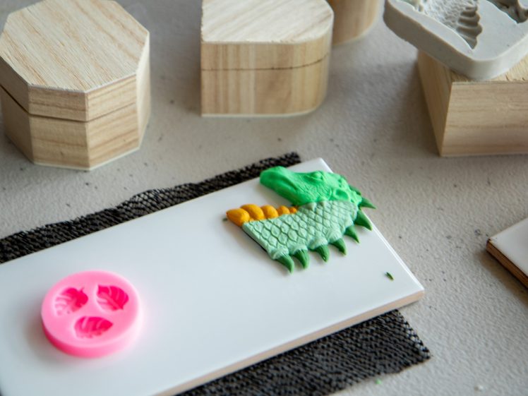Image of A green dragon head made from clay or fondant sits on a white rectangular tile, with a pink silicone mould nearby and wooden geometric blocks in the background.