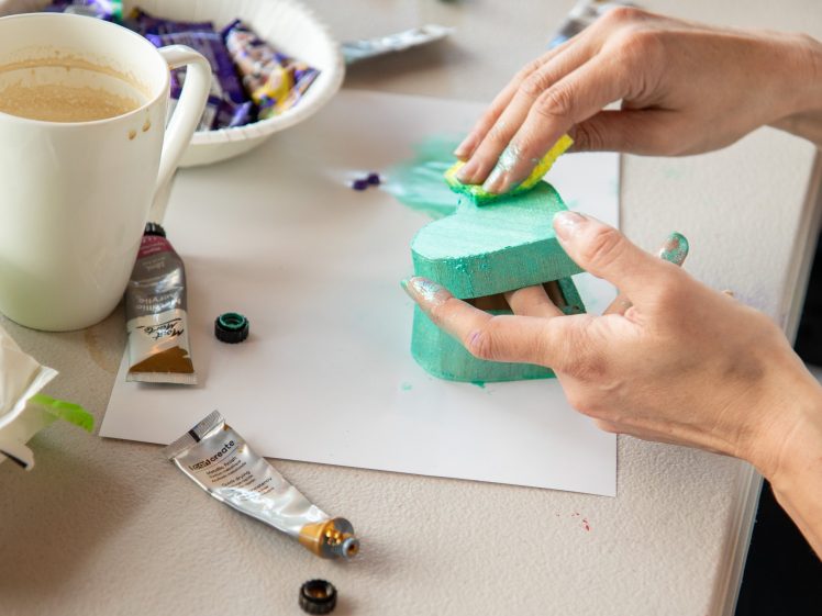 Image of A person paints a turquoise foam heart shape with yellow paint using their fingers. Paint tubes, a mug, and sweet wrappers are on the table beside them.