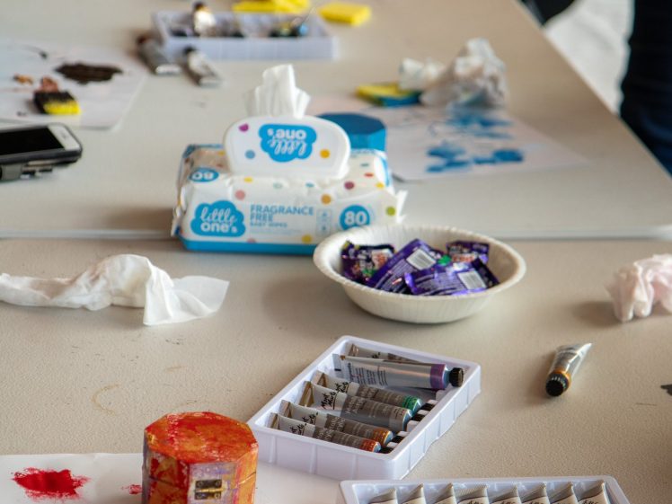 Image of A tabletop covered with art supplies, including tubes of paint, paint-stained sponges, paper with paint marks, a bowl with small packets, baby wipes, and tissues. Some people are partially visible in the background.