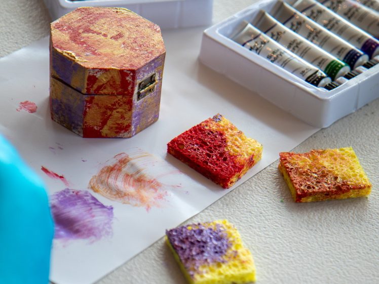 Image of A painted hexagonal box, colourful sponges, and trays of paint tubes are on a table. A gloved hand is visible in the lower left corner, and some paint marks are on the white sheet under the items.