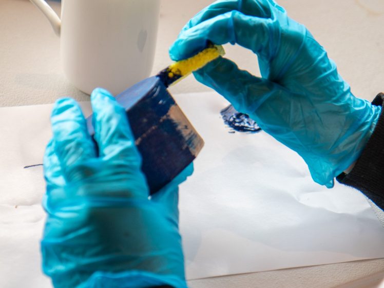 Image of A person wearing blue gloves paints a small blue object with a yellow brush, whilst a white mug with coffee sits on the table nearby.