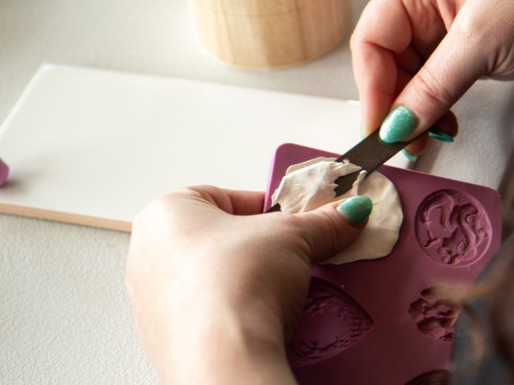 Image of A person with green painted nails uses a metal tool to press white clay into a purple silicone mould, with a round wooden object and white tile nearby on a table.