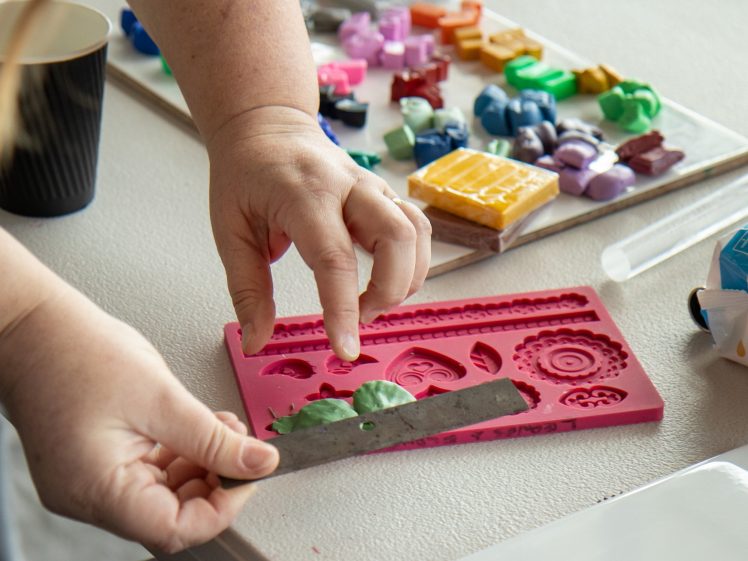 Image of A person uses a metal tool to work with green clay in a pink silicone mould, with colourful moulds and crafting materials on a table nearby.
