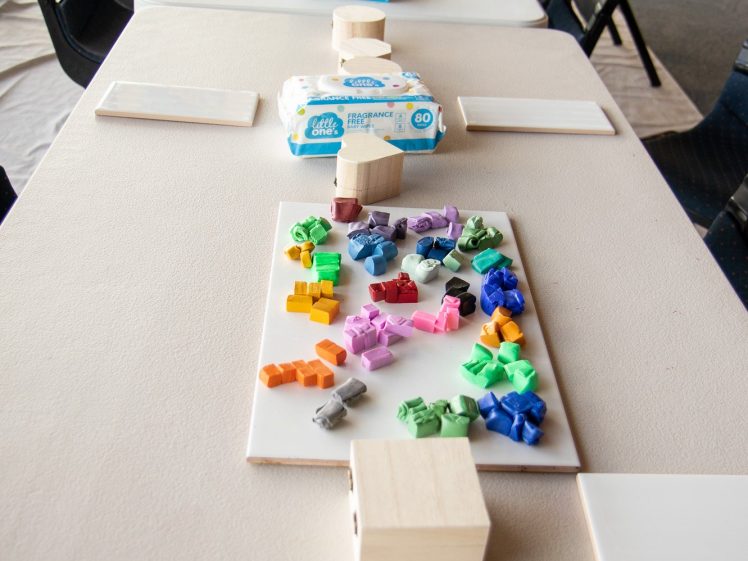 Image of A long table set up for a craft activity with blank canvases, colourful crayons, tissue boxes, and small wooden blocks. Black chairs are arranged on both sides of the table in a bright room.