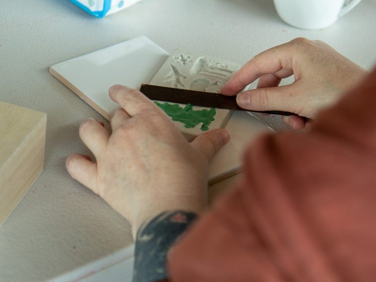 Image of A person uses a tool to spread green paint onto a white ceramic tile with raised designs, working carefully on a table. A mug and other materials are visible in the background.