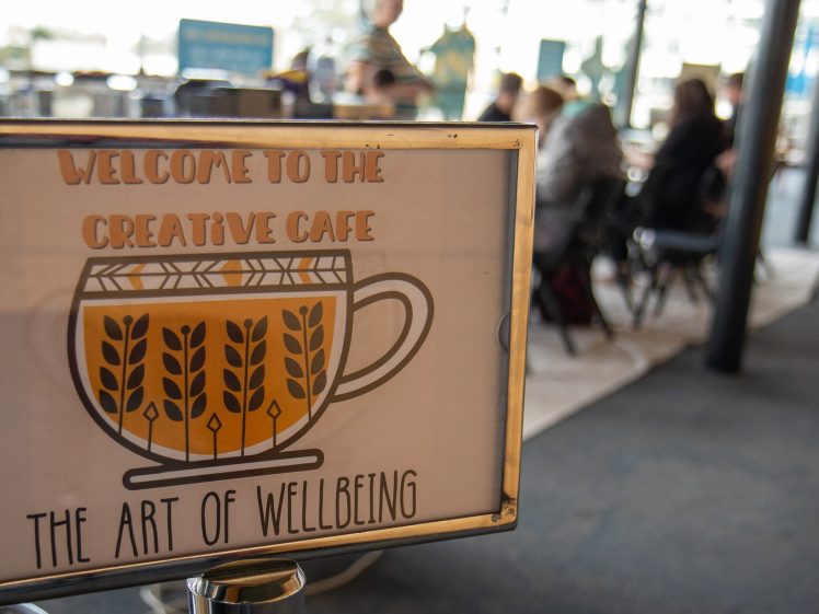Image of A sign reading Welcome to the Creative Café: The Art of Wellbeing with a cup illustration stands in focus, whilst people are seated and talking in a blurred café setting in the background.