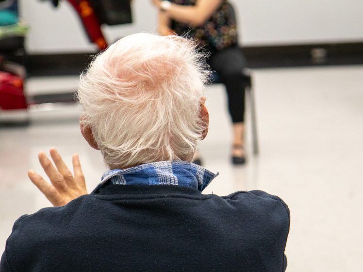 Image of An older man with white hair sits facing a woman who is gesturing with her hands in conversation. The scene takes place indoors, with both people seated and a red mobility scooter visible in the background.