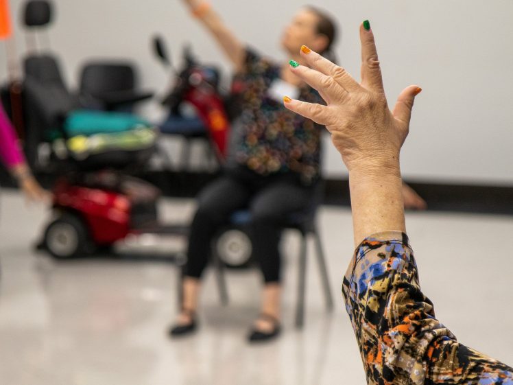 Image of A person sits on a chair with one arm raised, fingers spread, in a spacious room. Another person's raised hand, also with spread fingers, is visible in the foreground. Both wear patterned clothing.