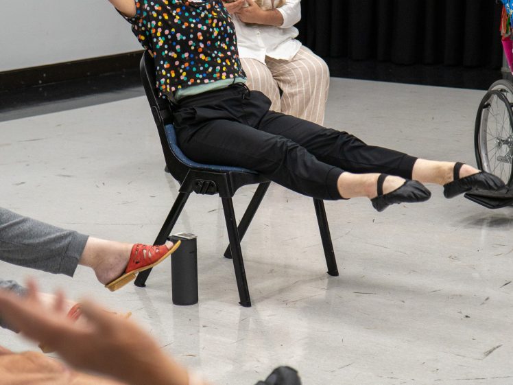 Image of A group of people participate in a seated dance class, raising their arms and legs. One woman in front stretches with arms extended and feet lifted, while another sits behind her, watching and holding a mobile.