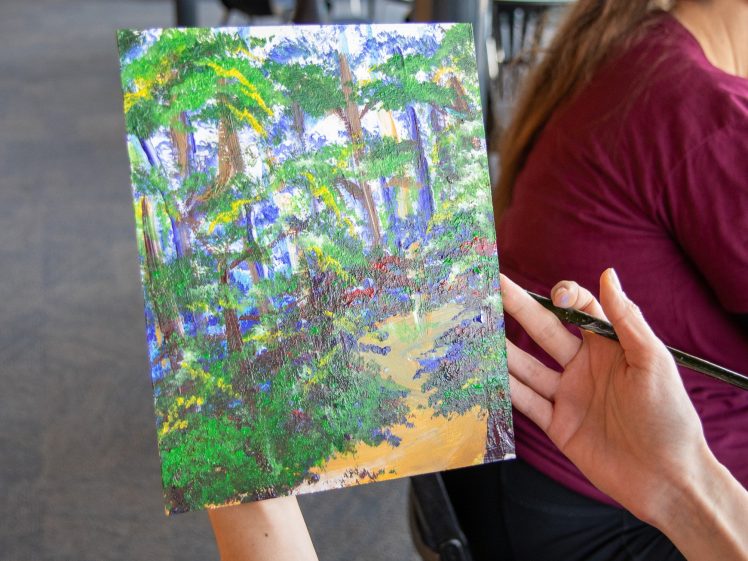 Image of A person holds a small canvas with a colourful painting of a forest scene, featuring green trees and a sunlit path, while holding a paintbrush in their other hand. The person wears a maroon shirt.