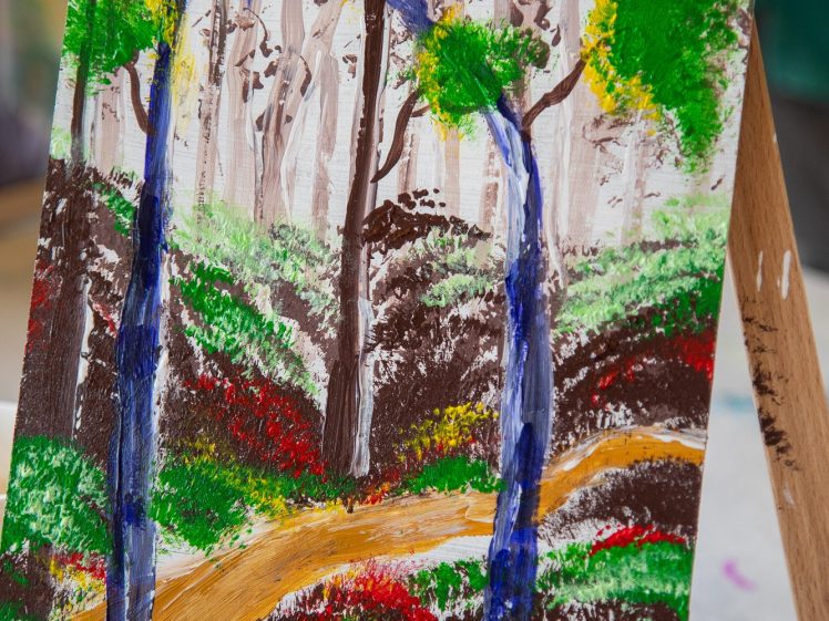 Image of A colourful painting on an easel shows a dirt path winding through a vibrant forest with green, yellow, and red foliage, brown tree trunks, and a background of tall, light trees.