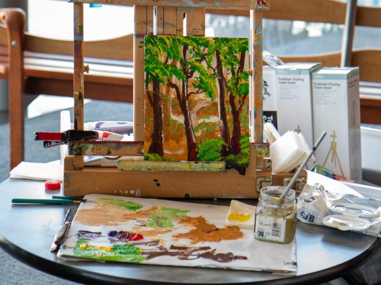 Image of A painting of a forest scene sits on an easel atop a round table, surrounded by paint tubes, brushes, a palette with mixed colours, and art supplies in a bright indoor setting.