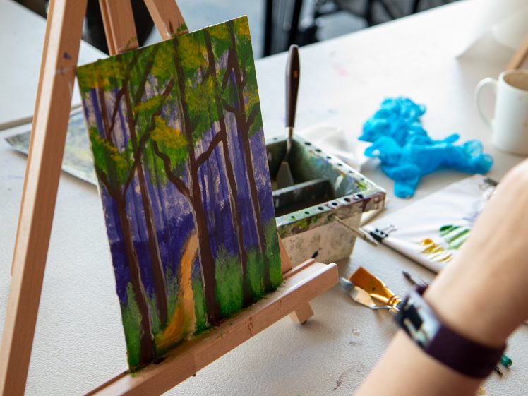 Image of A small painting of a forest with a path, placed on an easel. Art materials, a palette knife, blue gloves, and a mug are on the table nearby. A person's arm wearing a dark watch is visible in the foreground.