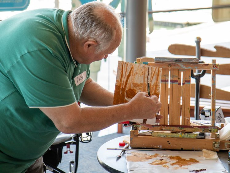 Image of An older man with grey hair, wearing a green polo shirt, paints on a small wooden easel indoors. Art supplies and paint are scattered on the table in front of him.