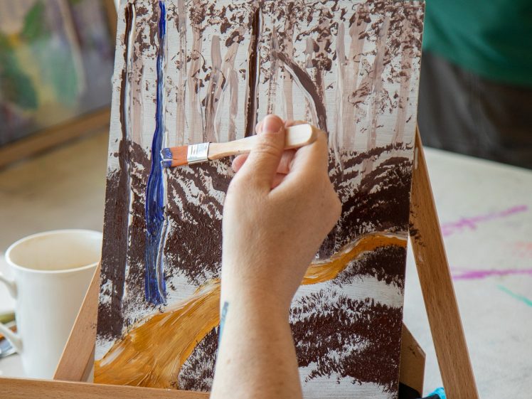 Image of A person paints a landscape of a forest scene with a yellow path on a canvas set on an easel. Their hand holds a brush adding blue to a tree. A white mug and blue gloves are on the table.