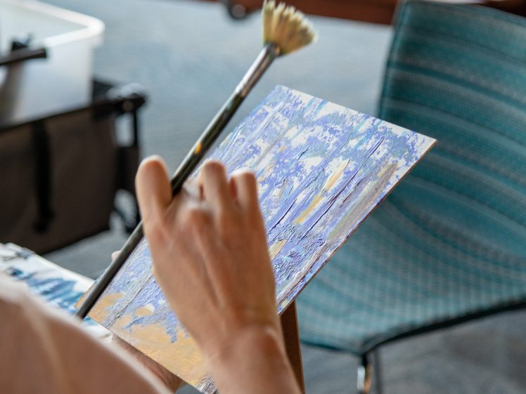 Image of A person holds a paintbrush and paints textured blue, white, and gold strokes on a small canvas. The scene appears indoors, with a chair and other art materials in the background.