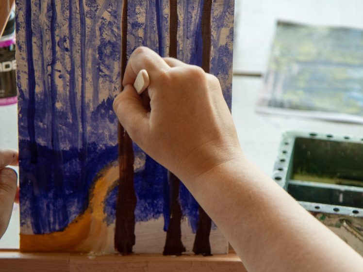 Image of A person uses white chalk or pastel to add details to a painting of a forest with tall trees and a winding path, set on an easel. Art materials and another painting are visible in the background.
