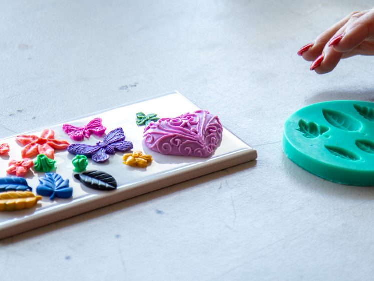 Image of A person shapes coloured clay or fondant using a green silicone mould with leaf patterns, while decorated butterflies, leaves, and heart shapes rest on a white tile nearby.