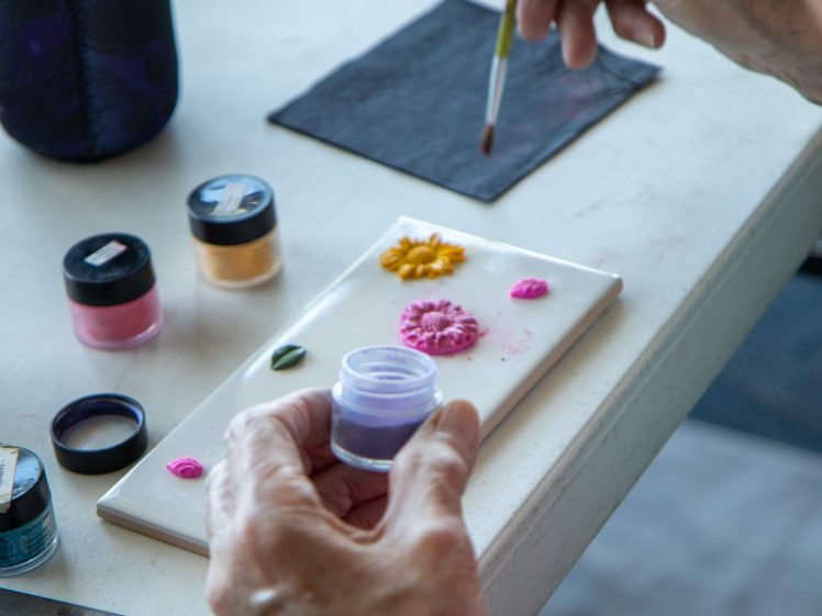 Image of A person paints decorative flowers in pastel colours on a small white tile using a fine brush, with open jars of paint and silicone moulds on a white table.