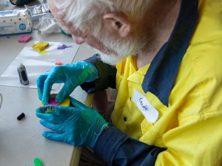 Image of An elderly man with white hair and beard, wearing a yellow and blue shirt and blue gloves, sits at a table crafting with colourful clay pieces. A name tag reading Cedric is on his shirt.
