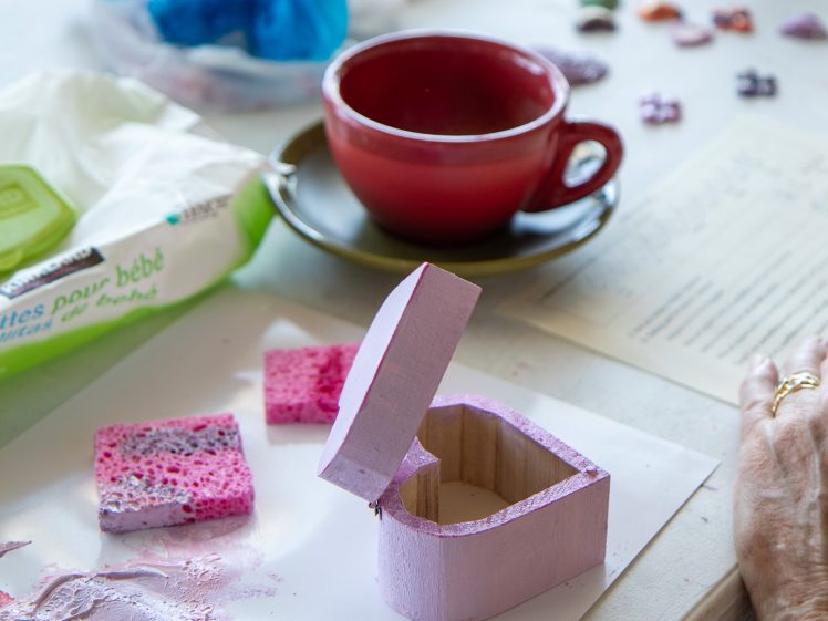 Image of A small, open, pink wooden box sits on a table with paint sponges, a red cup and saucer, baby wipes, watercolours, and scattered craft supplies around it. A hand rests nearby on a piece of paper.