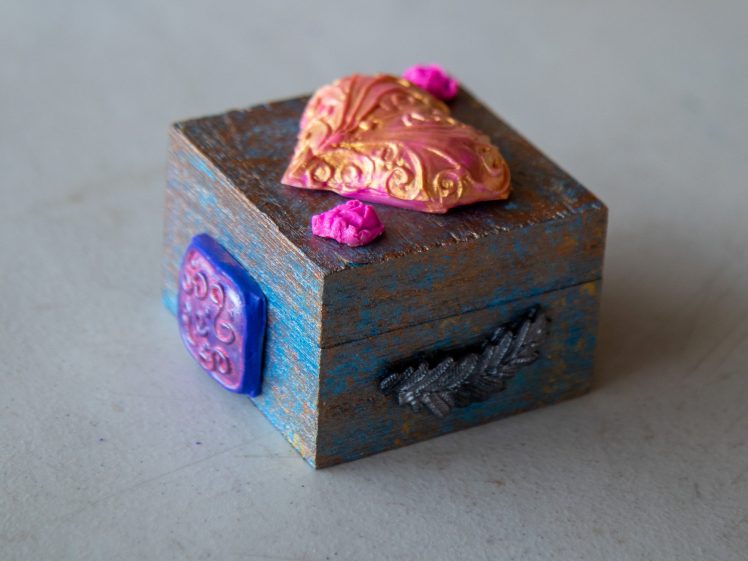 Image of A small, rustic wooden box with a weathered blue finish, decorated with a gold and pink heart, pink roses on top, a purple decorative side accent, and a silver leaf pattern on the front.