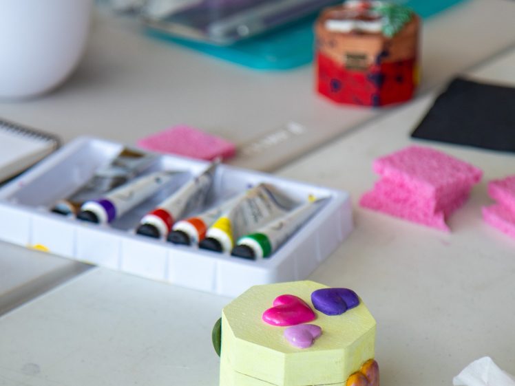 Image of A hexagonal yellow box decorated with colourful heart shapes sits on a table with paint tubes, pink sponges, art supplies, and a tissue nearby.