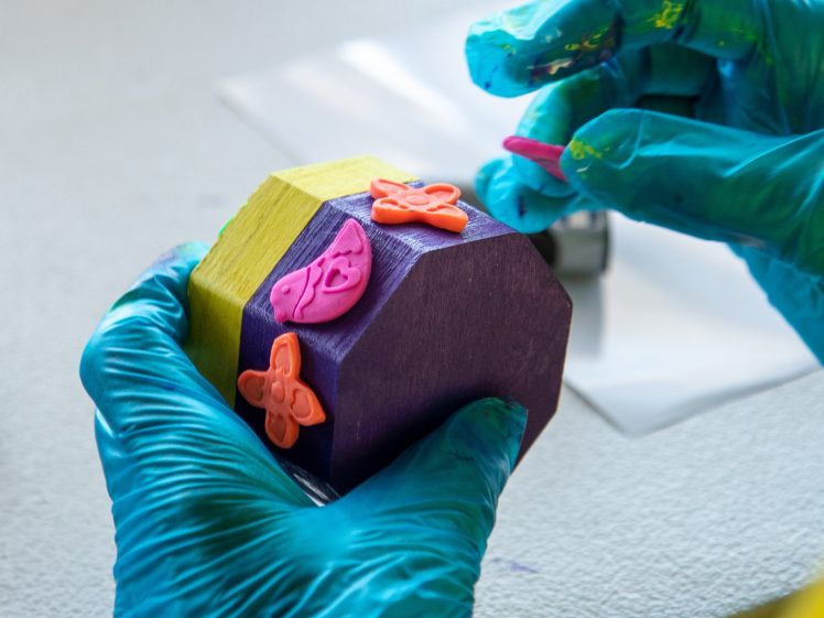 Image of A person wearing blue gloves decorates a wooden block painted yellow and purple with small, pink rubber shapes. The block is held in one hand whilst the other hand presses a shape onto its surface.