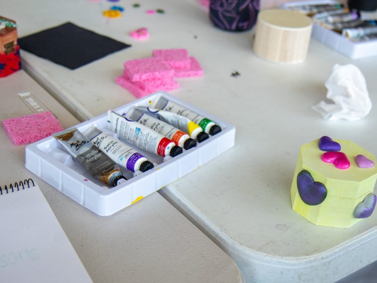 Image of A table with art supplies including paint tubes, pink sponges, a tissue, a notepad, and a small hexagonal box decorated with colourful heart shapes.