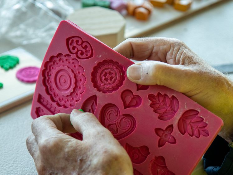 Image of A person’s hands hold a pink silicone mould with various decorative shapes. In the background, colourful moulded clay pieces are arranged on a table.