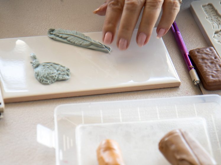 Image of A person's hand works with clay, shaping a leaf and a flower design on a white tile. Nearby are a pencil, a clay tool, and two pieces of brown clay on a clear tray.