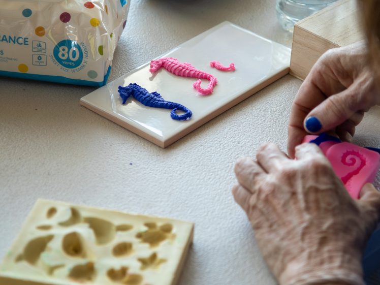 Image of A person crafts colourful seahorse shapes using silicone moulds and clay. Finished pink and blue seahorses are displayed on a white tile, with supplies and a packet of wipes nearby.