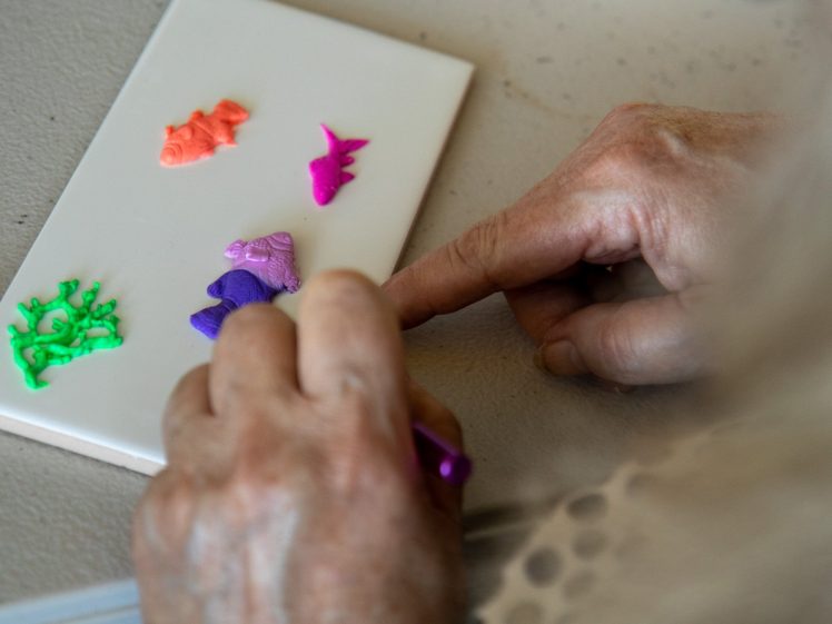 Image of A person uses their fingers to arrange small, colourful clay fish shapes on a white tile. The fish are orange, pink, green, and purple. The person's hands are in focus, with the rest of the image slightly blurred.