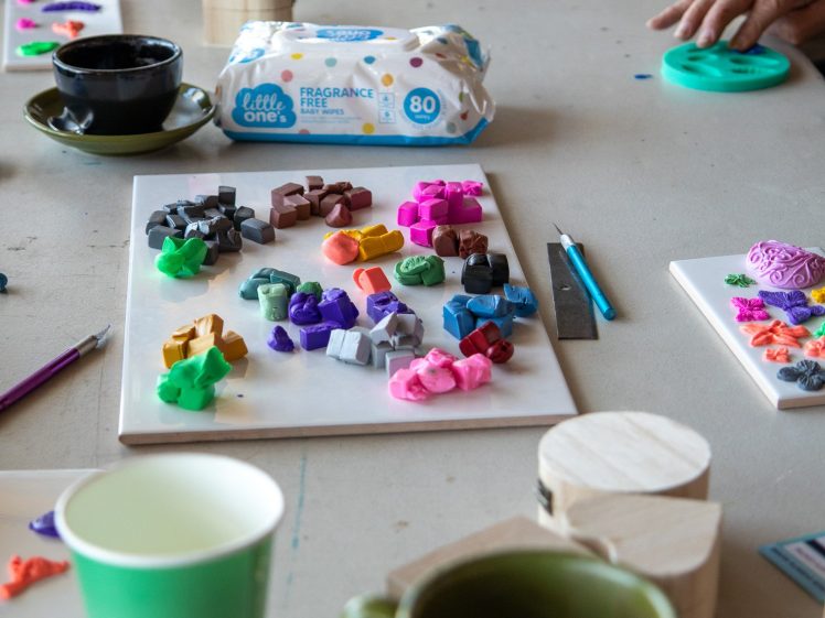 Image of A table with colourful clay creations, cups, wipes, wooden blocks, and crafting tools. Several people’s hands are visible as they work on their projects. The scene suggests a group craft activity.