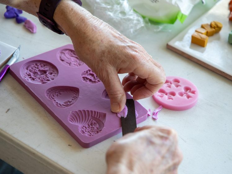 Image of A person uses a knife to shape purple modelling clay, pressing it into a pink silicone mould with various shell designs on a table with other clay tools and materials.