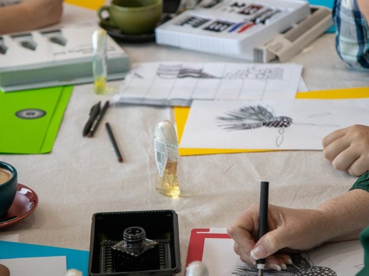 Image of People sitting around a table practising ink drawing with pens and paper. Art supplies, notebooks, and cups are scattered on the table. The focus is on hands drawing and various art materials.