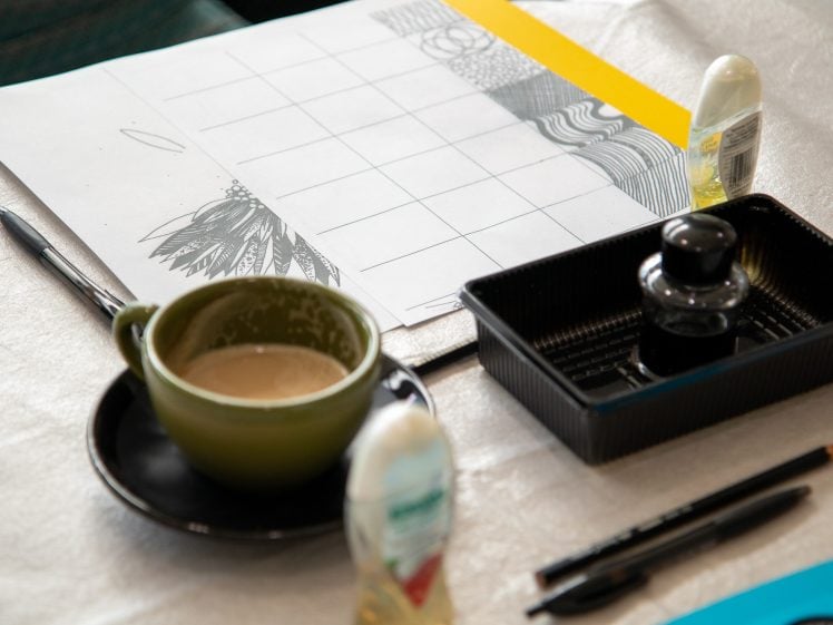 Image of A green coffee cup with foam sits on a saucer next to a sheet of paper with a grid and black patterns, pens, a pencil, correction fluid, and a black ink bottle on a table.