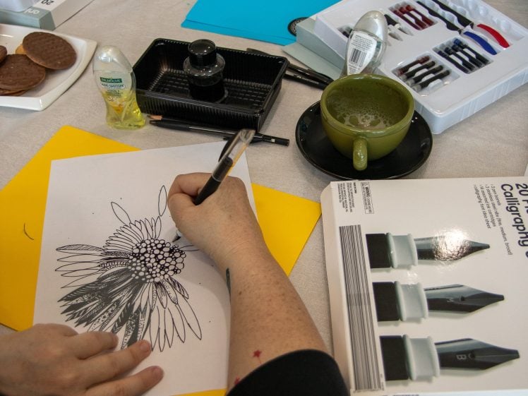Image of A person draws a detailed flower design on white paper with a black pen. Art supplies, coloured pens, a green coffee cup, biscuits, and a yellow folder are on the table.